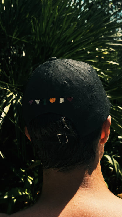 Back view of black NoMuss cap with colorful embroidered flag detail and adjustable metal clasp, worn outdoors against tropical greenery
