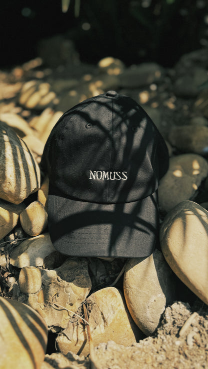 Black NoMuss branded cap with white embroidered logo, resting on sun-dappled river rocks in natural light