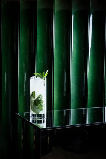 Een verfrissende Nomuss Mojito in een highball glas met crushed ice, verse muntblaadjes en een rietje, geserveerd op een donkere bar met groene glanzende tegels op de achtergrond.